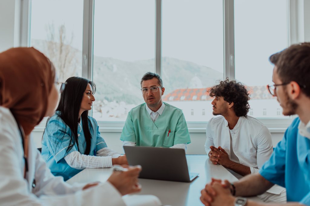 A group of multiethnic medical professionals including doctors, surgeons, and nurses are gathered in a hospital setting discussing patient care and using modern technology to address challenges in the medical field